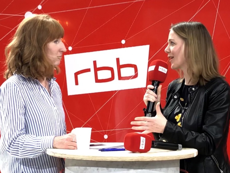 Cathleen holding a microphone by rbb news, answering the moderator's question, who is standing in front of her.